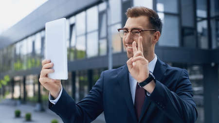 happy businessman in glasses having video call while showing peace sign and holding digital tablet outsideの写真素材