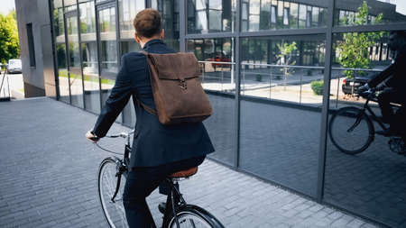 back view of businessman in suit with leather brown backpack riding bicycle near building