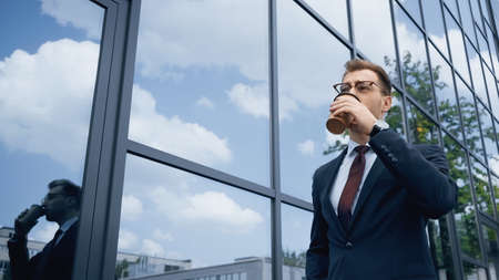 businessman in glasses drinking coffee to go near building with glass facadeの写真素材