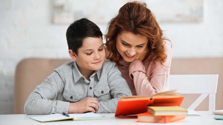 Smiling woman sitting near preteen son and books on blurred foreground