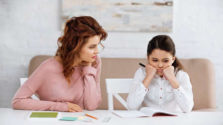Thoughtful girl sitting near notebook, digital tablet and mother at homeの写真素材
