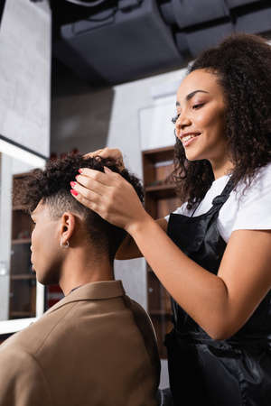 Smiling african american hairdresser looking at hair of man in salonの写真素材