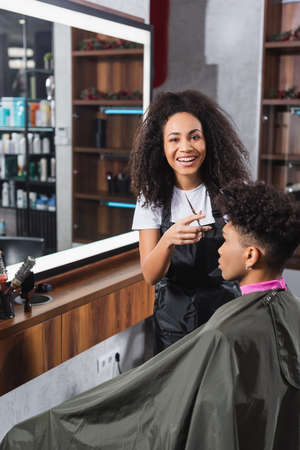 African american hairstylist with scissors smiling at camera near client in salonの写真素材
