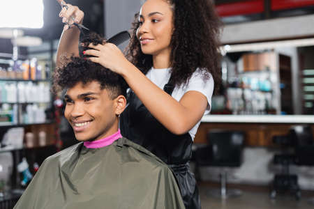 Cheerful african american client sitting near hairdresser holding scissors and comb in salonの写真素材