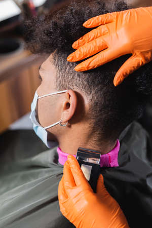Hairstylist in latex gloves trimming neck of african american man in medical mask and capeの写真素材