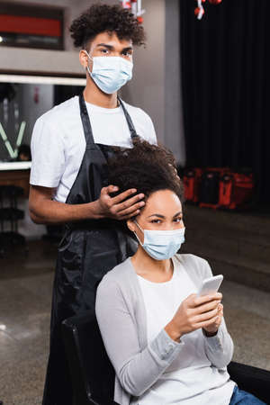 African american woman with smartphone looking at camera near hairstylist in medical mask