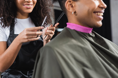 Cropped view of comb and scissors in hands of african american hairstylist near smiling client on blurred foregroundの写真素材