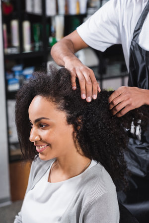 Cheerful african american woman sitting near hairdresser in salonの写真素材