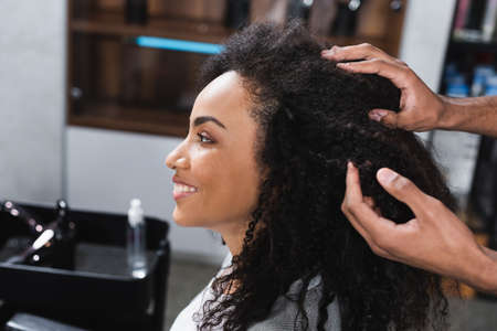 Side view of smiling african american client sitting near hairstylist touching hairの写真素材