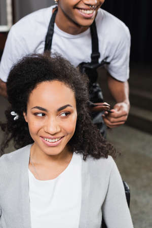 Smiling african american woman looking away near hairstylist with hair straightener on blurred backgroundの写真素材