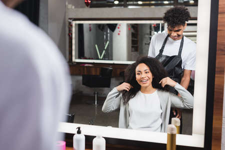 Cheerful hairstylist in apron looking at african american woman touching hairの写真素材