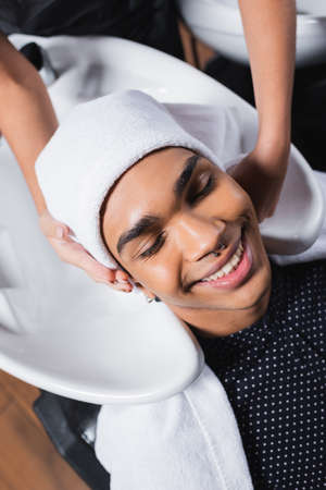 Top view of smiling african american client sitting near sink and hairstylist with towel