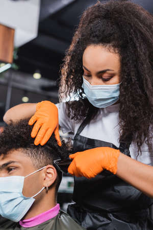 African american hairdresser in medical mask and gloves trimming hair of client on blurred foregroundの写真素材