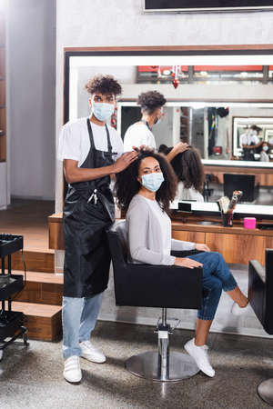 African american hairstylist in protective mask looking at camera near young womanの写真素材