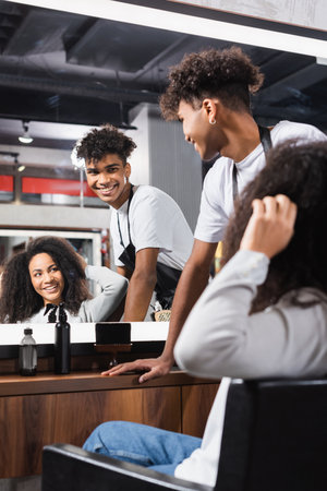 Smiling african american hairstylist looking at mirror near woman touching hair on blurred foregroundの写真素材