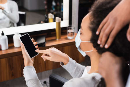 African american client in medical mask pointing at smartphone near hairdresser on blurred foregroundの写真素材