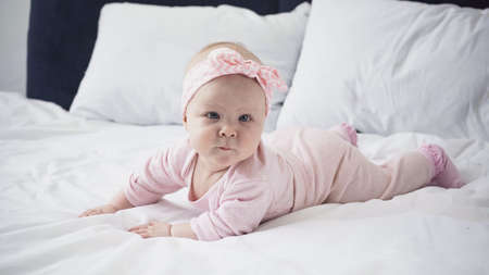infant baby girl in headband with bow lying on bed with white bedding