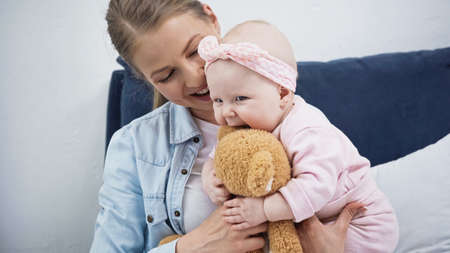 happy woman holding soft toy near infant daughter in headband with bow