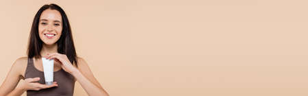 young woman smiling at camera while holding cosmetic cream isolated on beige, bannerの写真素材