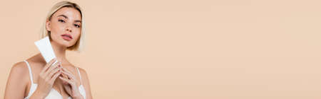 blonde woman looking at camera while holding cosmetic cream isolated on beige, bannerの写真素材