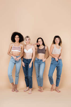 full length view of young multiethnic women in bras and jeans posing on beigeの写真素材