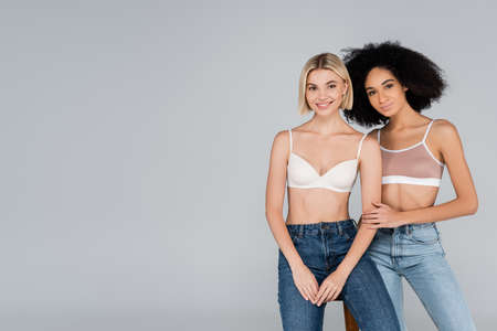 young interracial women in bras and jeans looking at camera isolated on grayの写真素材