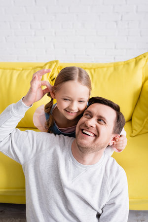 cheerful girl and laughing father touching ears of each other while having fun at homeの写真素材