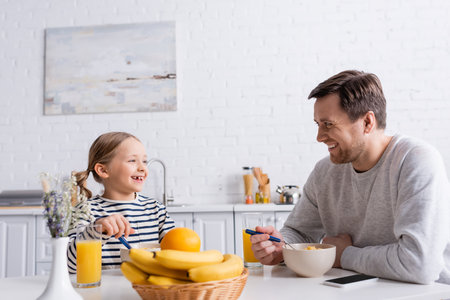 father and daughter smiling at each other while eating corn flakes near fresh fruits on blurred foregroundの写真素材