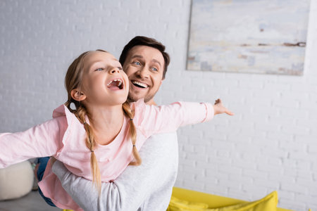 happy man holding amazed daughter showing fly gesture while having fun at homeの写真素材