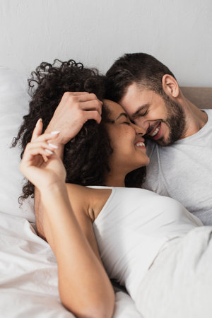 cheerful multiethnic man and woman hugging while lying in bedの写真素材