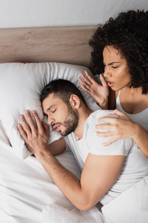 top view of displeased african american woman looking at bearded man sleeping on bed