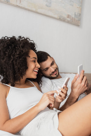 cheerful interracial couple using cellphones in bedroomの写真素材