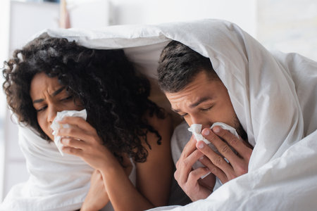 interracial sick couple sneezing in napkins under blanketの写真素材
