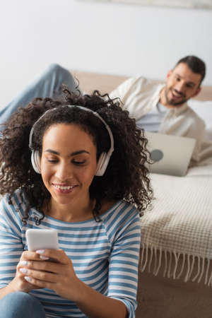 smiling african american woman in wireless headphones holding smartphone near boyfriend using laptop on blurred backgroundの写真素材