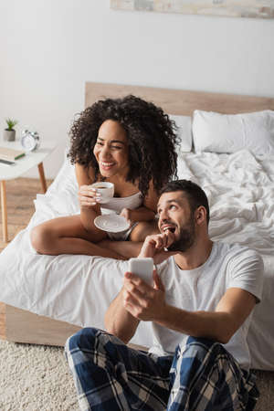 happy african american woman holding cup with coffee while bearded boyfriend using smartphone in bedroomの写真素材