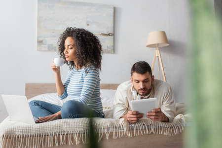 african american woman sitting on bed, holding cup and using laptop near boyfriend with digital...
