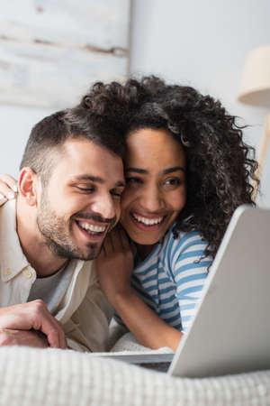 positive interracial couple chilling on bed and watching movie on laptopの写真素材