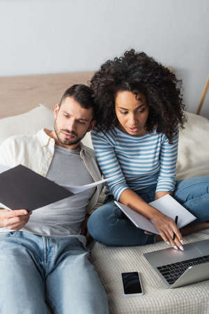 african american woman holding notebook near boyfriend and looking at laptop