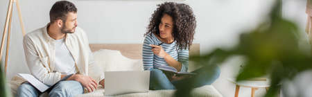 african american woman pointing at boyfriend sitting with folder near laptop on bed, banner