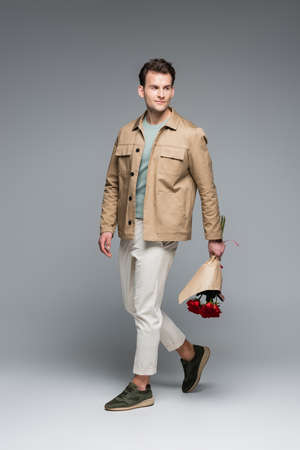 full length of trendy man holding bouquet of red roses wrapped in paper on gray
