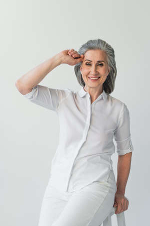 Mature woman in white clothes smiling at camera near chair isolated on gray