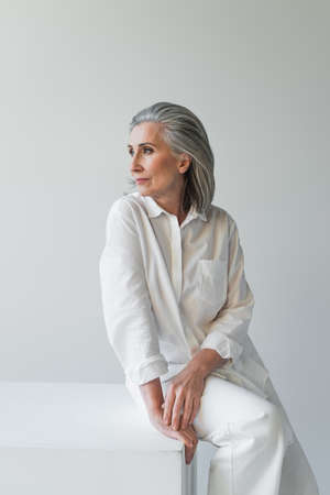 Mature woman sitting on white cube isolated on gray