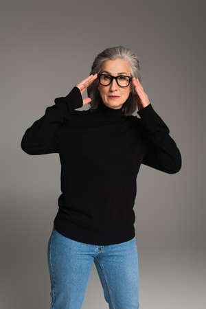 Mature woman in jumper, jeans and eyeglasses looking at camera isolated on grayの写真素材