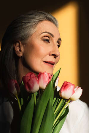 Middle aged woman holding pink tulips on yellow background with shadow