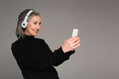 Smiling mature woman in headphones using smartphone isolated on grayの写真素材