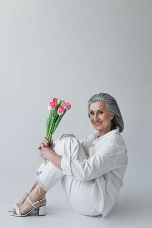 Middle aged woman holding flowers and smiling at camera while sitting on gray backgroundの写真素材
