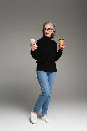 Mature woman using smartphone and holding takeaway drink on gray backgroundの写真素材