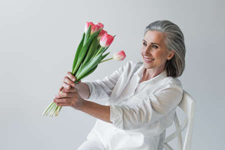 Mature woman smiling while looking at tulips on chair isolated on grayの写真素材