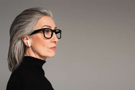 Mature woman in turtleneck, eyeglasses and earphone isolated on gray