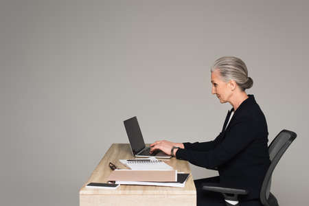 Side view of businesswoman using laptop near papers isolated on gray